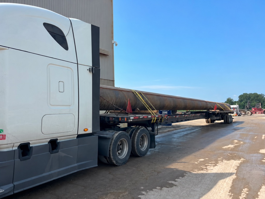 White tractor and long flatbed carrying a pipe in an industrial yard